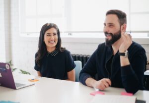 A smiling woman with a black collared blouse sitting next to a bearded man who is laughing 