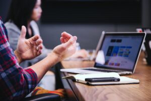 A man in a plaid shirt gestures during a meeting, while a woman looks introspectively at her computer screen