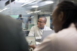  Man wearing glasses, framed by other people in the meeting room, smiles at something being said